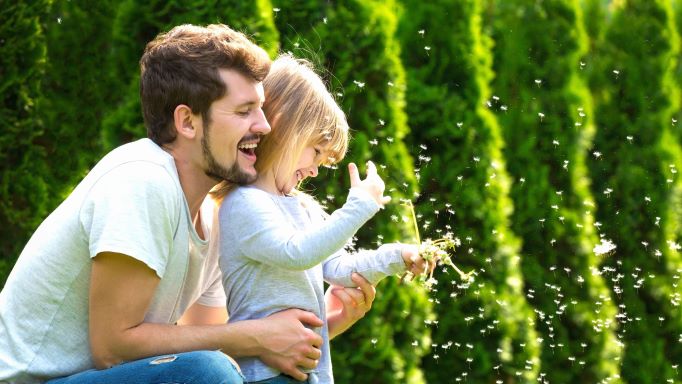 Otec s malou dcerou se smějí a dcera si hraje s odkvetlou pampeliškou, za nimi jsou zelené stromy.